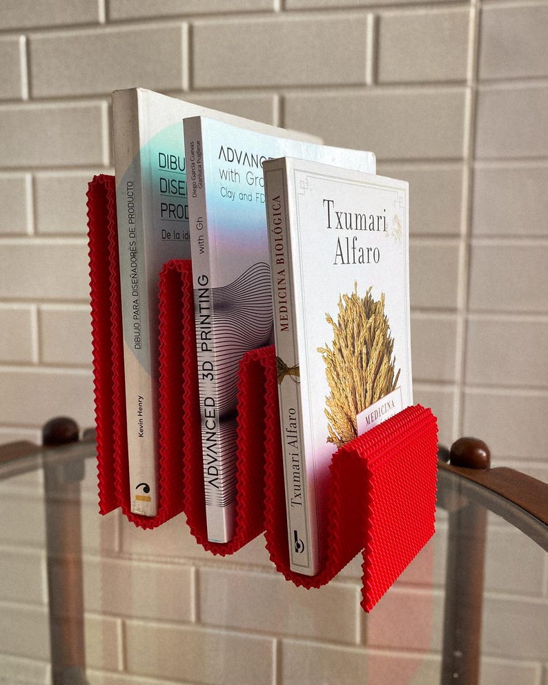 May include: A red, textured book holder with three books. The books have white covers and titles in Spanish and English. The holder is made of a flexible material and is designed to hold books upright. The books are on a glass surface.