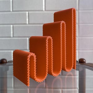 May include: An abstract art piece featuring four orange, textured, folded shapes with a zigzag edge. The shapes are arranged in a stepped pattern, with a light orange glow. The background is a white brick wall.