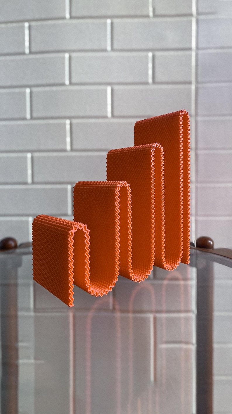 May include: An abstract art piece featuring four orange, textured, folded shapes with a zigzag edge. The shapes are arranged in a stepped pattern, with a light orange glow. The background is a white brick wall.