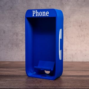 Op de afbeelding: Een blauwe telefooncelvormige telefoonstandaard met het woord "Phone" erop gedrukt. De standaard heeft een gleuf voor een telefoon en een uitsparing voor de oplaadpoort.
