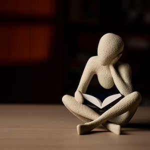 Könnte beinhalten: Beigefarbene Figur einer Person, die im Schneidersitz ein Buch liest. Der Kopf der Figur ruht auf ihrer Hand, was auf Nachdenklichkeit hindeutet. Das Buch ist geöffnet, und der Hintergrund ist dunkel.