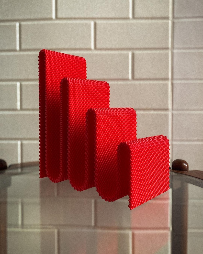 May include: Four red, textured, rectangular objects of varying heights are arranged in a stepped pattern. The objects have a woven appearance and are set against a neutral, tiled background. The objects are reflected on a glass surface.