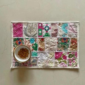 May include: A rectangular, patchwork placemat with a variety of colorful, embroidered fabric squares. A small, white bowl with a gold rim holds a serving of almonds. The placemat has a white border.