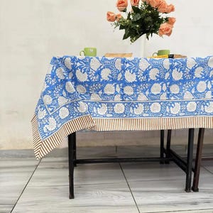 Peut inclure: Une nappe rectangulaire ornée d'un motif floral bleu et blanc imprimé au bloc, avec une bordure rayée marron et blanche. Le tissu recouvre une table à structure métallique noire, surmontée d'un bouquet de roses pêche et deux tasses vertes.