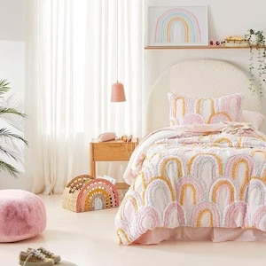 Pode incluir: Uma cena de quarto com uma cama com um edredom e uma almofada com padrão de arco-íris. Uma cesta em forma de arco-íris a condizer está no chão, juntamente com uma otomana rosa. Uma impressão de arte de arco-íris está pendurada acima da cama.
