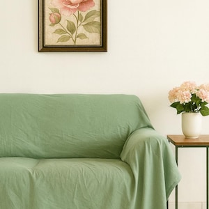 Può includere: Un copridivano in tessuto verde salvia drappeggiato su un divano, posizionato accanto a un tavolino con un vaso bianco contenente fiori rosa chiaro, e una stampa botanica incorniciata di una grande peonia rosa appesa alla parete.