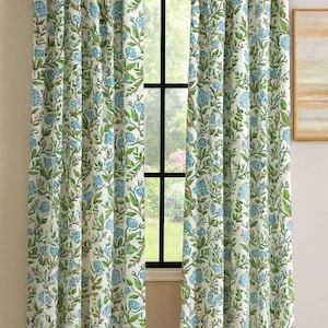 May include: Floral patterned curtains in a light cream color. The curtains feature a repeating pattern of blue flowers and green leaves on vines. The curtains are hanging in front of a window, with a view of the outside visible through the glass.