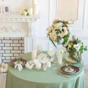 May include: A round table covered with a sage green tablecloth, set for a formal occasion. The table is adorned with floral arrangements, candles, and place settings. A white fireplace and floral wreath are in the background.
