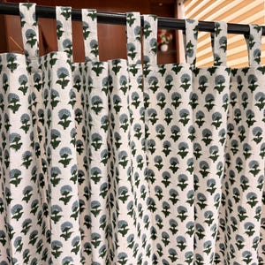 May include: A tab-top curtain featuring an off-white fabric base with a repeating block-print pattern of dark green stems and light blue, rounded, fan-shaped floral motifs. The curtain hangs from a black rod with evenly spaced fabric loops.