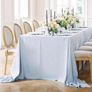 May include: A light blue tablecloth covers a long dining table, set for a formal event. White plates, glassware, and floral arrangements decorate the table. Gold-framed chairs with cream seats are arranged around the table.