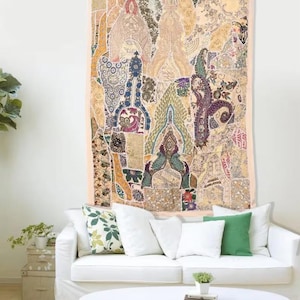 Puede incluir: Un gran tapiz rectangular con un diseño de patchwork en tonos beige, marrón, verde y azul cuelga de una pared blanca. El tapiz presenta patrones y texturas intrincados. Debajo, un sofá blanco con almohadas y una mesa de centro redonda blanca completan la escena.