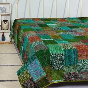 Op de afbeelding: Een patchwork sprei bestaande uit diverse groene, blauwgroene en veelkleurige stoffen vierkanten met complexe bloem- en geometrische patronen. De sprei is voorzien van zichtbare stikseldetails die een gestructureerd oppervlak creëren.