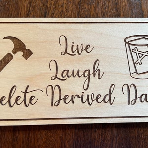 May include: A wooden sign with the text "Live Laugh Delete Derived Data" in a cursive font. The sign also features a hammer and a glass with a card inside.