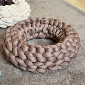Peut inclure: Un lit pour animaux de compagnie rond et tressé, de couleur taupe clair. Le lit est fait d'un matériau épais et doux et présente un motif tressé. Il est posé sur un tapis de couleur neutre. Un pot tressé blanc et beige est en arrière-plan.