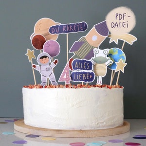 Könnte beinhalten: Ein weißer Kuchen mit Weltraum-Dekorationen. Die Dekorationen zeigen einen Astronauten, Planeten, eine Rakete und einen Frosch. Auf den Dekorationen steht "DU RAKETE!" und "ALLES LIEBE". Der Kuchen steht auf einem Holzsockel.