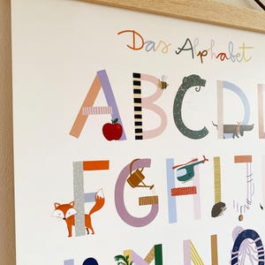 May include: A colorful alphabet poster with the words "Das Alphabet" at the top. Each letter is decorated with illustrations of objects and animals, including an apple, bee, fox, and carrot. The poster is framed with a light wood.
