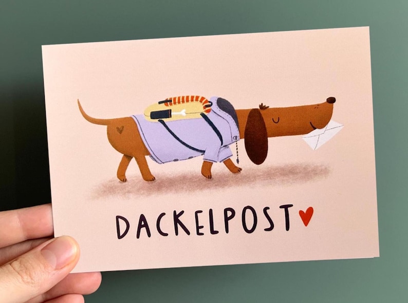 May include: A rectangular greeting card with a light pink background featuring a brown dachshund wearing a light purple jacket and a yellow backpack, carrying a white envelope in its mouth. The text DACKELPOST is printed in black with a red heart.
