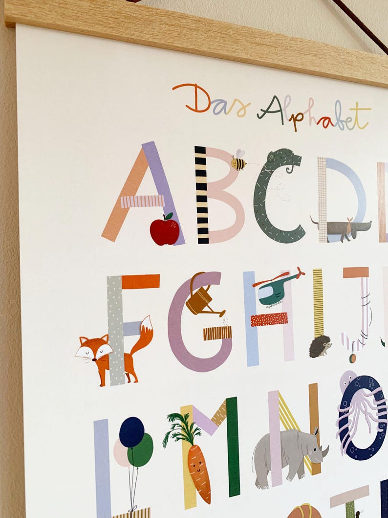 May include: A colorful alphabet poster with the words "Das Alphabet" at the top. Each letter is decorated with illustrations of objects and animals, including an apple, bee, fox, and carrot. The poster is framed with a light wood.
