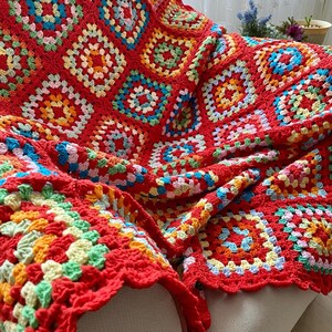 Peut inclure: Une couverture en crochet rouge avec un motif de carr&eacute;s de grand-m&egrave;re color&eacute;s. La couverture est faite de plusieurs couleurs, dont le rouge, l'orange, le jaune, le vert, le bleu et le rose.