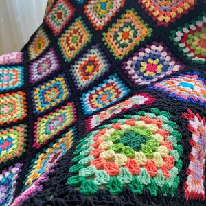 Può includere: Una coperta all'uncinetto colorata con sfondo nero e un motivo a quadrati della nonna. La coperta è realizzata con una varietà di colori, tra cui rosso, arancione, giallo, verde, blu, viola e rosa.