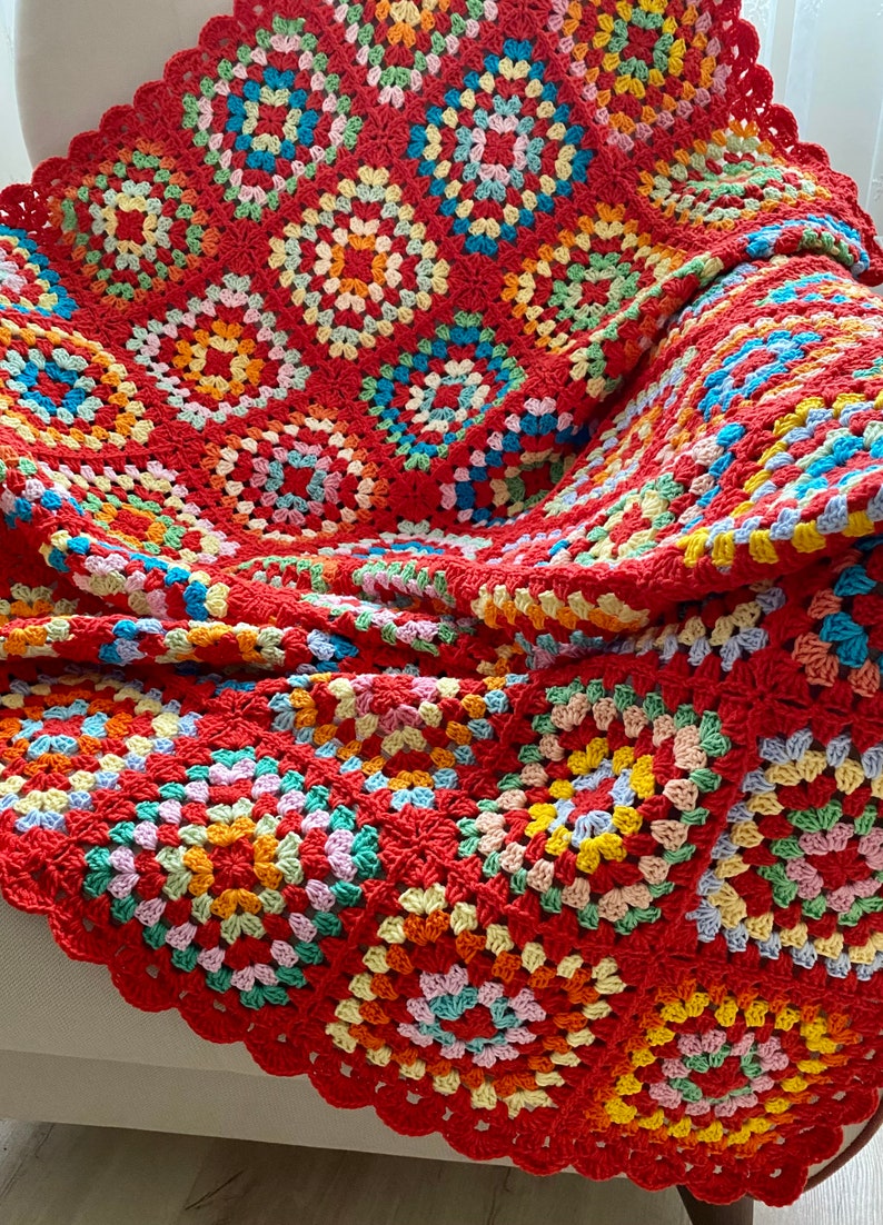 Peut inclure: Une couverture en crochet color&eacute;e avec un fond rouge et des carr&eacute;s de grand-m&egrave;re multicolores. La couverture est pli&eacute;e et drap&eacute;e sur un fauteuil blanc.