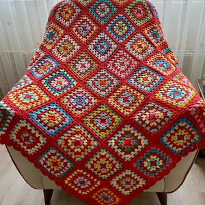 Peut inclure: Une couverture en crochet color&eacute;e avec un fond rouge et des carr&eacute;s de grand-m&egrave;re multicolores. La couverture est drap&eacute; sur un fauteuil blanc.