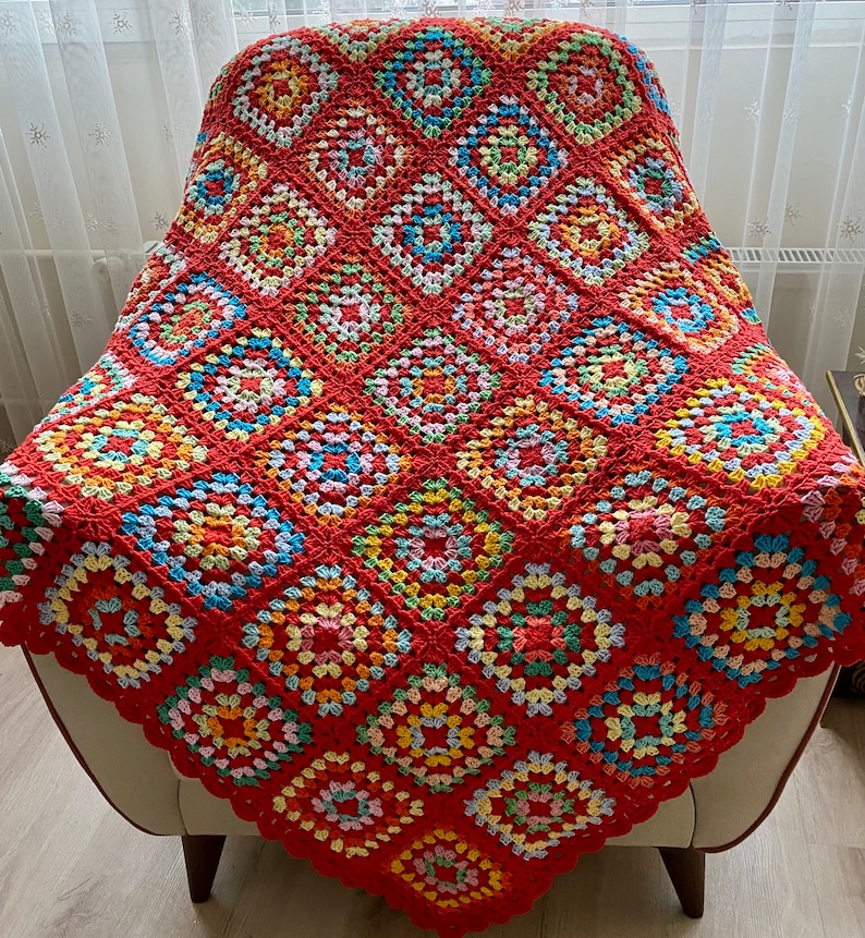 Peut inclure: Une couverture en crochet color&eacute;e avec un fond rouge et des carr&eacute;s de grand-m&egrave;re multicolores. La couverture est drap&eacute; sur un fauteuil blanc.