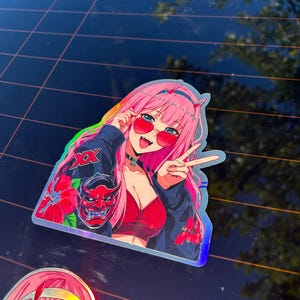 May include: A holographic vinyl sticker featuring an anime character with long pink hair, red heart-shaped sunglasses, and a black jacket. The character is making a peace sign, wearing a red top, and is decorated with red hibiscus flowers and a red oni mask.