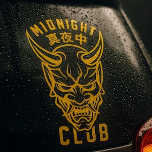 May include: A gold-colored decal featuring a horned demon face with the words "MIDNIGHT 真夜中 CLUB" in gold lettering. The decal is applied to a black surface, with water droplets visible.
