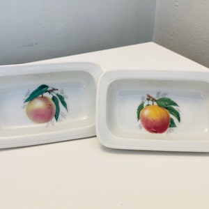 May include: Two white ceramic butter dishes with a raised rim. Each dish features a hand-painted design of a single peach with green leaves.