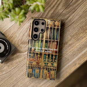 May include: A phone case featuring a library design with a gold gate and shelves filled with books. The case has the text "I get all my books from the restricted section". The case is on a wooden surface.