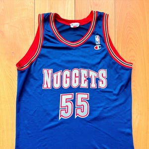 Può includere: Una canotta da basket blu dei Nuggets Champion con dettagli rossi e bianchi. La canotta presenta la scritta "Nuggets" in bianco con un contorno argentato e il numero "55" in bianco con un contorno rosso. È visibile un logo NBA.