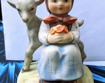 Puede incluir: Una figurita de porcelana de una niña con un vestido azul y blanco que sostiene una cesta de flores. Está sentada junto a una cabra blanca.