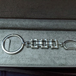 Könnte beinhalten: Ein silberner Schlüsselanhänger mit einem Kettenglied-Design und einem silbernen Ring. Der Schlüsselanhänger hat an einem Ende einen silbernen Haken.
