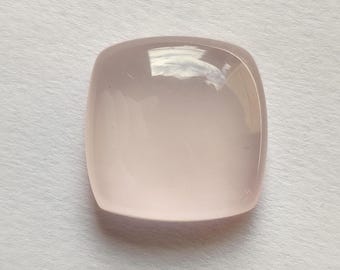 May include: A polished, pale pink gemstone with a rounded square shape. The stone has a smooth, reflective surface, and the colour is a soft, pale pink. The gemstone is likely a cabochon, suitable for jewellery making or collecting.