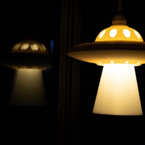 May include: A yellow, UFO-shaped pendant light with a white, cone-shaped shade, emitting a warm glow. The design features circular cutouts on the top. A reflection of the light is visible against the dark background. A quirky lighting fixture.