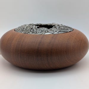 May include: A wooden bowl with a dark brown finish and a silver-coloured metal rim with a floral design. The bowl has a rounded, oval shape. The rim is decorated with a detailed floral pattern. A coin is visible in the background.
