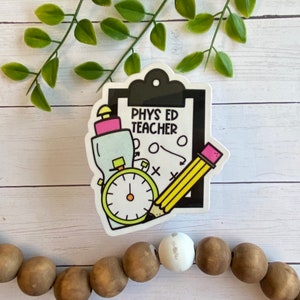 May include: A white sticker with a black and white illustration of a clipboard with the text "PHYS ED TEACHER" on it. The clipboard has a water bottle, a stopwatch, and a pencil on it.