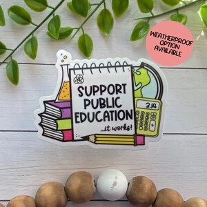 Puede incluir: Una pegatina blanca con una ilustraci&oacute;n colorida de una pila de libros, un l&aacute;piz, una calculadora, una probeta, un globo terr&aacute;queo y una libreta que dice "Apoyar la educaci&oacute;n p&uacute;blica... &iexcl;funciona!" La pegatina est&aacute; sobre una superficie de madera.