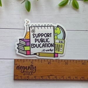 Puede incluir: Una pegatina blanca con una ilustraci&oacute;n colorida de un cuaderno, una calculadora, un globo terr&aacute;queo, un l&aacute;piz y una pila de libros. El cuaderno dice "Apoya la educaci&oacute;n p&uacute;blica... &iexcl;funciona!"