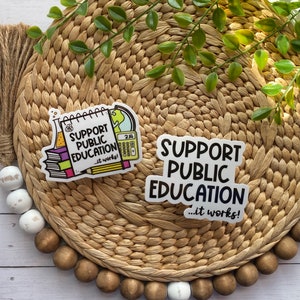 Puede incluir: Dos pegatinas con el texto "Apoyar la educaci&oacute;n p&uacute;blica... &iexcl;funciona!" Una pegatina presenta una ilustraci&oacute;n de dibujos animados de un cuaderno, una calculadora, una probeta y un globo terr&aacute;queo. La otra pegatina es una simple pegatina blanca con texto negro.