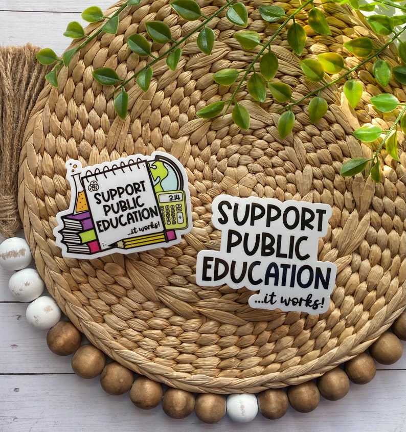 Puede incluir: Dos pegatinas con el texto "Apoyar la educaci&oacute;n p&uacute;blica... &iexcl;funciona!" Una pegatina presenta una ilustraci&oacute;n de dibujos animados de un cuaderno, una calculadora, una probeta y un globo terr&aacute;queo. La otra pegatina es una simple pegatina blanca con texto negro.