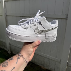 May include: White low-top sneaker with a jeweled Nike swoosh and accents. The shoe features white laces, a white sole, and the word "AIR" on the side. The sneaker is held up against a white fence.