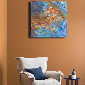 May include: Abstract painting with blue, brown, and gold colors. The painting is on a canvas and is hanging on a wall in a living room setting. A white armchair with a blue pillow is in the foreground.