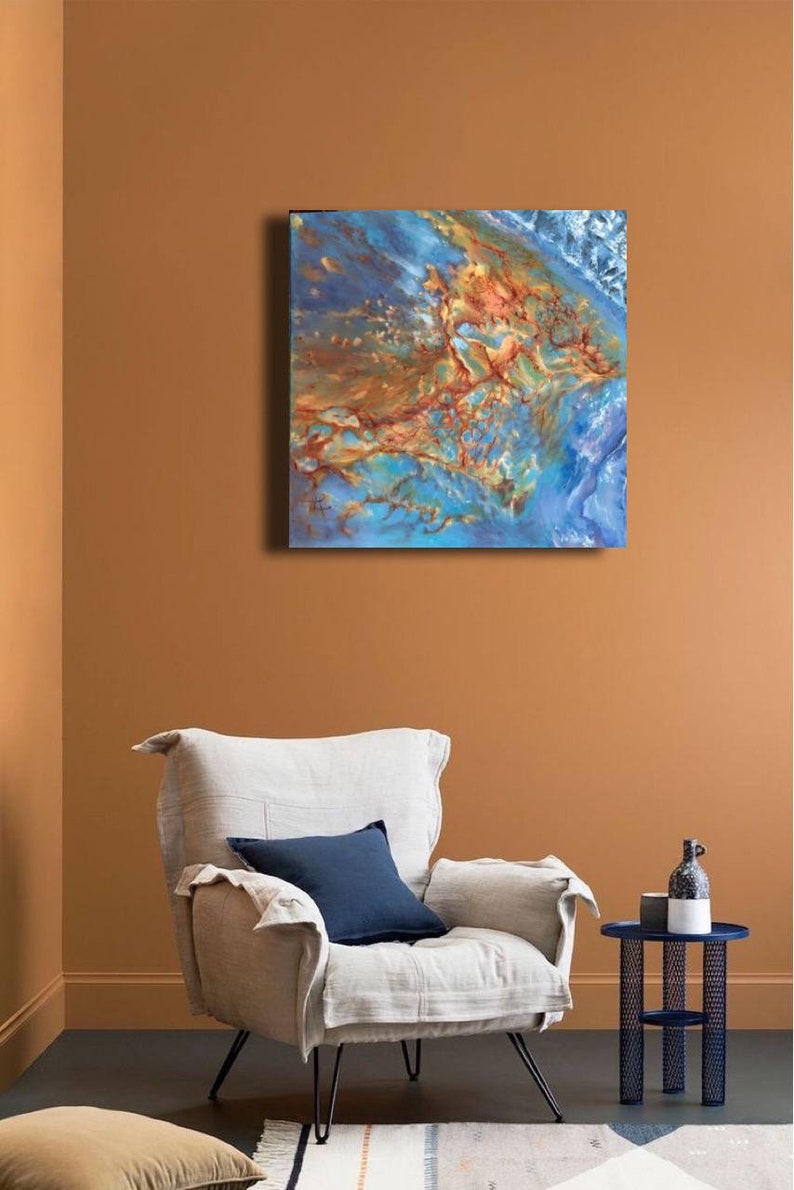May include: Abstract painting with blue, brown, and gold colors. The painting is on a canvas and is hanging on a wall in a living room setting. A white armchair with a blue pillow is in the foreground.
