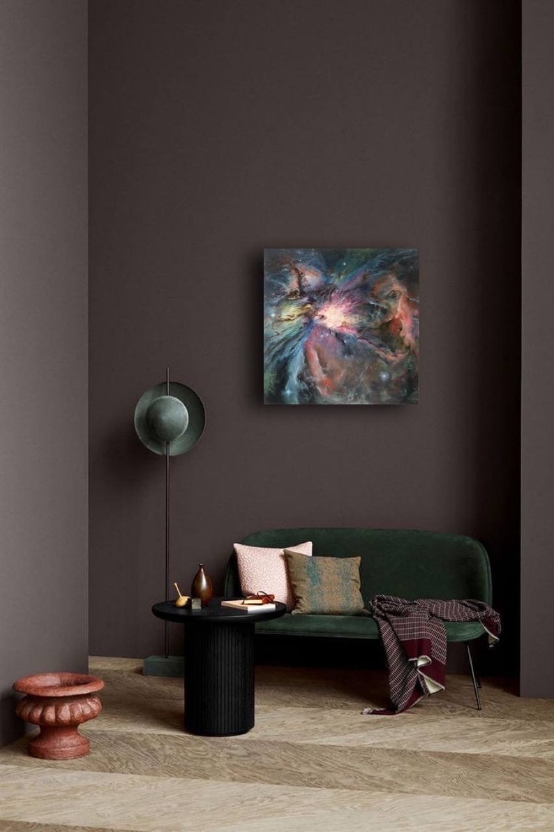 May include: A green velvet sofa with a throw blanket and pillows in a living room setting. A painting of a colorful nebula hangs on the wall above the sofa. A black side table with a vase and a book sits in front of the sofa.
