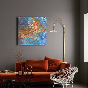 May include: Abstract painting with blue, orange, and brown colors. The painting is hanging on a gray wall above a burnt orange sofa and a white wire chair.