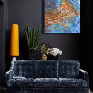 May include: A dark blue velvet sofa with a tufted design sits in a room with a dark gray wall. A large abstract painting with blue, brown, and gold colors hangs above the sofa. A tall yellow lamp and a potted plant are also in the room.