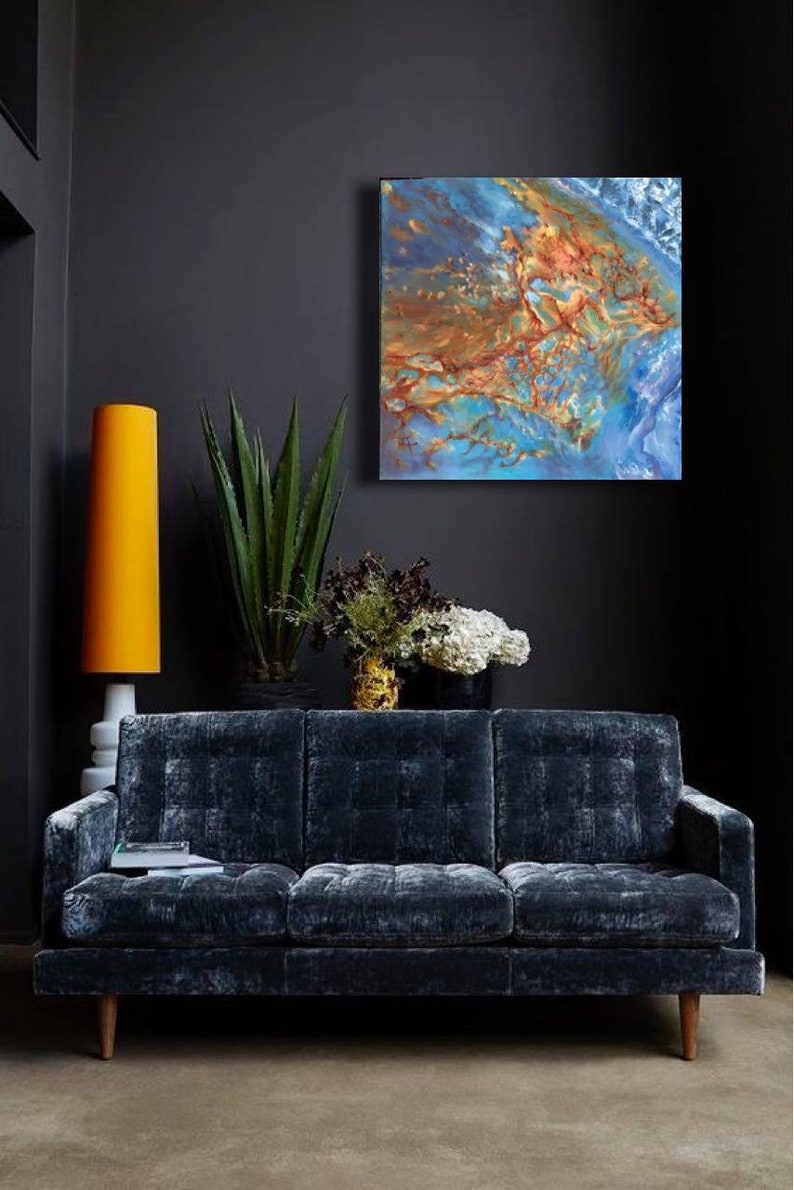 May include: A dark blue velvet sofa with a tufted design sits in a room with a dark gray wall. A large abstract painting with blue, brown, and gold colors hangs above the sofa. A tall yellow lamp and a potted plant are also in the room.