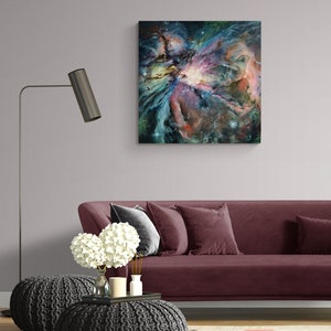 May include: A painting of a nebula in shades of blue, purple, pink, and green. The nebula is surrounded by a dark blue background with white stars. The painting is framed and hanging on a wall above a burgundy sofa.